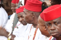 A group of men wearing traditional attire featuring red hats and white clothing. They appear to be in a formal or ceremonial setting, and some of them have necklaces made of beads. The men are sitting closely together, engaged in conversation or focused on an event.