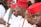 A group of men wearing traditional attire featuring red hats and white clothing. They appear to be in a formal or ceremonial setting, and some of them have necklaces made of beads. The men are sitting closely together, engaged in conversation or focused on an event.
