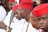 A group of men wearing traditional attire featuring red hats and white clothing. They appear to be in a formal or ceremonial setting, and some of them have necklaces made of beads. The men are sitting closely together, engaged in conversation or focused on an event.