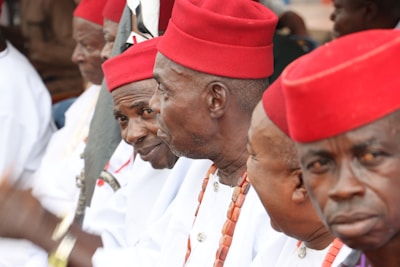A group of men wearing traditional attire featuring red hats and white clothing. They appear to be in a formal or ceremonial setting, and some of them have necklaces made of beads. The men are sitting closely together, engaged in conversation or focused on an event.