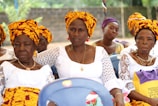 A group of women are sitting together, dressed in vibrant traditional attire. They are wearing white lace dresses paired with colorful yellow and orange headscarves. The background is an outdoor setting with greenery and a concrete wall.