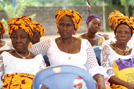 A group of women are sitting together, dressed in vibrant traditional attire. They are wearing white lace dresses paired with colorful yellow and orange headscarves. The background is an outdoor setting with greenery and a concrete wall.