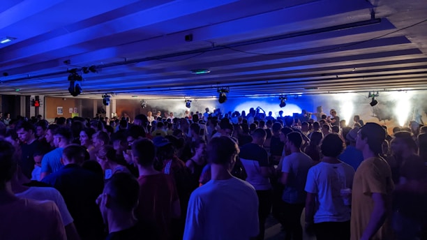 A crowded indoor party with numerous people gathered under a ceiling with blue lights and fog effects creating an energetic atmosphere. Most individuals are standing, enjoying the lively ambiance.