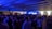 A crowded indoor party with numerous people gathered under a ceiling with blue lights and fog effects creating an energetic atmosphere. Most individuals are standing, enjoying the lively ambiance.