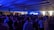 A crowded indoor party with numerous people gathered under a ceiling with blue lights and fog effects creating an energetic atmosphere. Most individuals are standing, enjoying the lively ambiance.