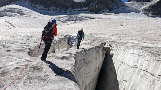 Two climbers equipped with helmets and backpacks navigate a snowy crevasse on a glacier. The vast expanse of ice and snow is surrounded by rocky mountains in the background. A trail of footprints can be seen leading towards the climbers.