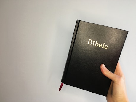 A hand is holding a black hardcover book with the word 'Bibele' written in gold lettering on the cover. The background is a plain, light-colored wall.