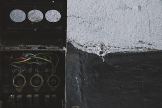 An old, open electrical junction box with visible wires in red, blue, yellow, and black. The box is mounted on a rough-textured wall, partially painted in black and white.