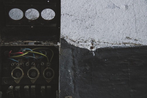 An old, open electrical junction box with visible wires in red, blue, yellow, and black. The box is mounted on a rough-textured wall, partially painted in black and white.