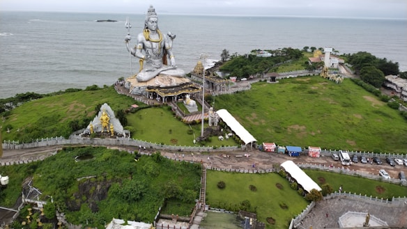 A large statue of a seated figure holding a trident overlooks a scenic landscape with lush greenery and ocean in the background. The statue appears to be a religious or cultural landmark surrounded by pathways and smaller structures.