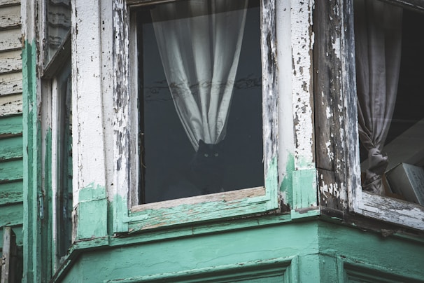 A close-up of a weathered, haunted mansion’s cracked window with a faint face peering out.
