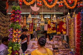 A vibrant market stall displays a wide array of colorful decorations, including garlands of flowers, golden hanging ornaments, and intricate craft items. Several people are browsing and shopping, surrounded by the lively and festive atmosphere.
