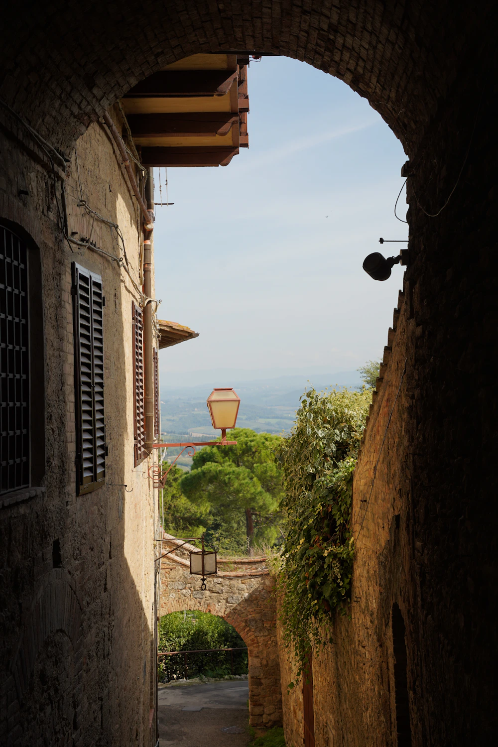 Doorkijk onder een stenen boog naar een smal straatje en het landschap buiten Siena