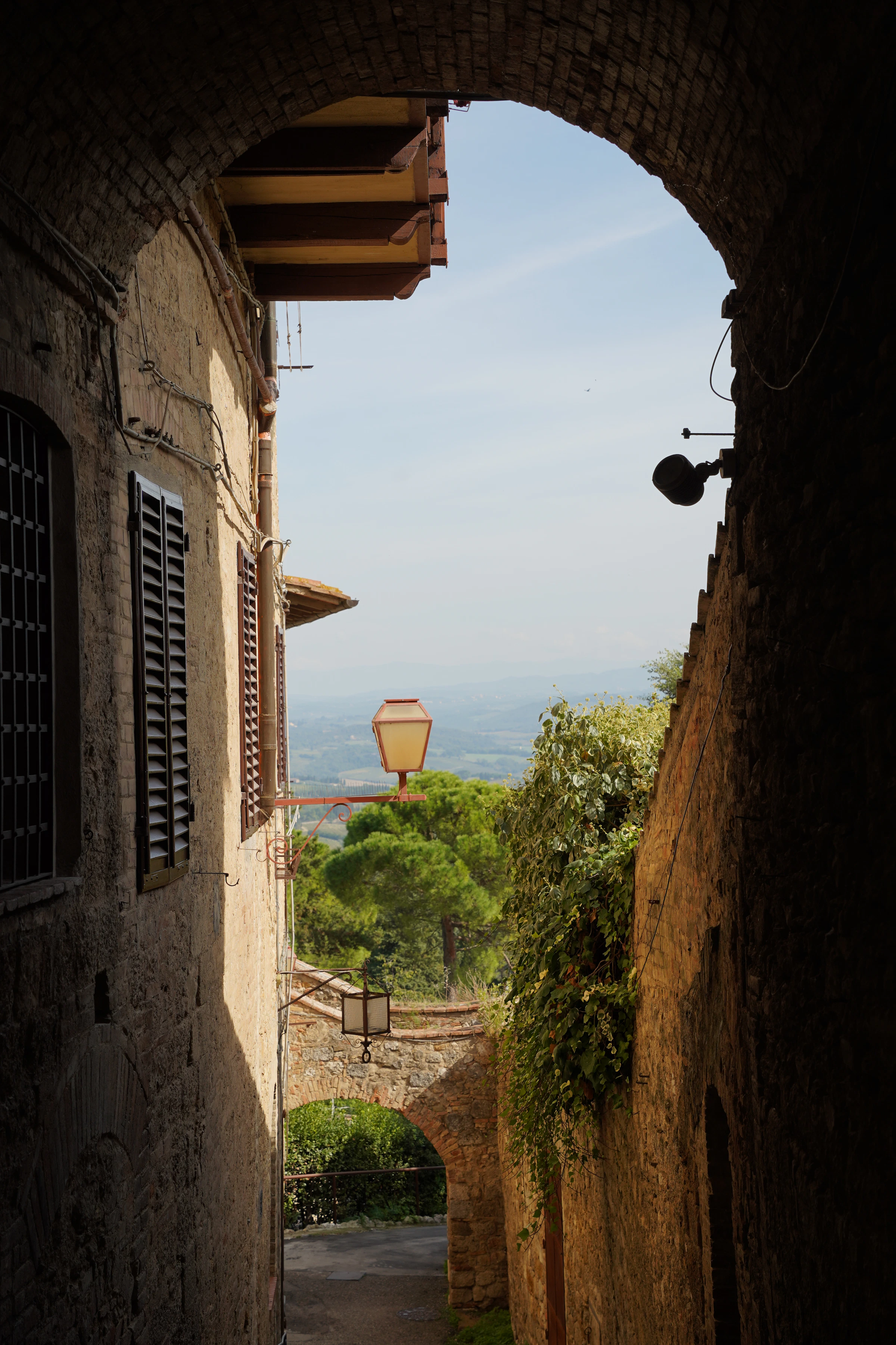 Doorkijk onder een stenen boog naar een smal straatje en het landschap buiten Siena
