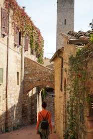 A narrow cobblestone street flanked by rustic stone buildings covered in ivy and vivid flowers. A person with a backpack walks along, framed by an archway and the tower of a historical structure in the background.