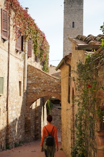 A narrow cobblestone street flanked by rustic stone buildings covered in ivy and vivid flowers. A person with a backpack walks along, framed by an archway and the tower of a historical structure in the background.