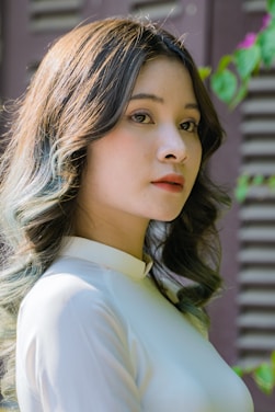 A woman with long wavy hair looks intently at the camera. She is wearing a light-colored traditional dress and is positioned against a background with wooden shutters and some greenery.