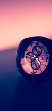 A wristwatch with a prominent large dial and multiple subdials, viewed at an angle. The watch face displays Roman numerals and reflective surfaces under a pinkish-purple ambient light.