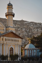 A mosque with an ornate minaret and a domed building beside it stand prominently. The structures feature intricate architectural details in soft pastel colors, surrounded by a fence. In the background, a hillside is covered with numerous small houses.