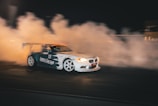 A drifting car emitting thick clouds of smoke as it maneuvers on a dark track. The vehicle is adorned with various decals and has a prominent rear spoiler, headlights illuminated. The environment suggests a high-speed, action-packed scene.