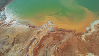 An aerial view of a landscape featuring a mix of green and orange hues spread across the surface, resembling a lake or wetland area. The contrasting colors suggest mineral deposits or algae presence. The terrain appears arid, with visible textures and patterns.