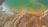 An aerial view of a landscape featuring a mix of green and orange hues spread across the surface, resembling a lake or wetland area. The contrasting colors suggest mineral deposits or algae presence. The terrain appears arid, with visible textures and patterns.