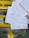 Several handwritten postcards being held in front of a yellow mailbox. The postcards contain messages in various languages and have barcodes at the bottom. The mailbox is mounted on a sidewalk alongside some posters.