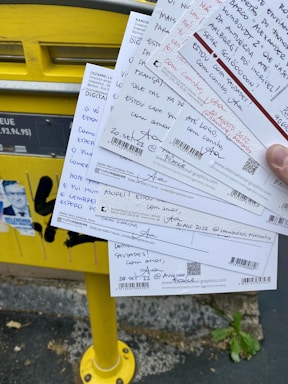 Several handwritten postcards being held in front of a yellow mailbox. The postcards contain messages in various languages and have barcodes at the bottom. The mailbox is mounted on a sidewalk alongside some posters.