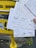 Several handwritten postcards being held in front of a yellow mailbox. The postcards contain messages in various languages and have barcodes at the bottom. The mailbox is mounted on a sidewalk alongside some posters.