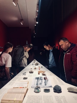 A group of people are gathered around a long, narrow table that displays various items such as rocks, papers, and different artifacts. The setting has a red backdrop with focused lighting on the objects, creating an intimate and contemplative atmosphere.