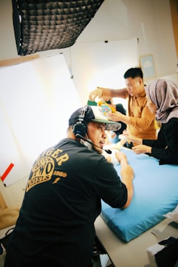 Three individuals are engaged in an activity indoors under professional lighting equipment. One person is wearing a black shirt and a cap with a headset, appearing to coordinate or give instructions. Another person in a brown shirt and a person wearing a light headscarf are manipulating objects on a blue cushioned surface. The room has a clean, organized setup with bright lighting.