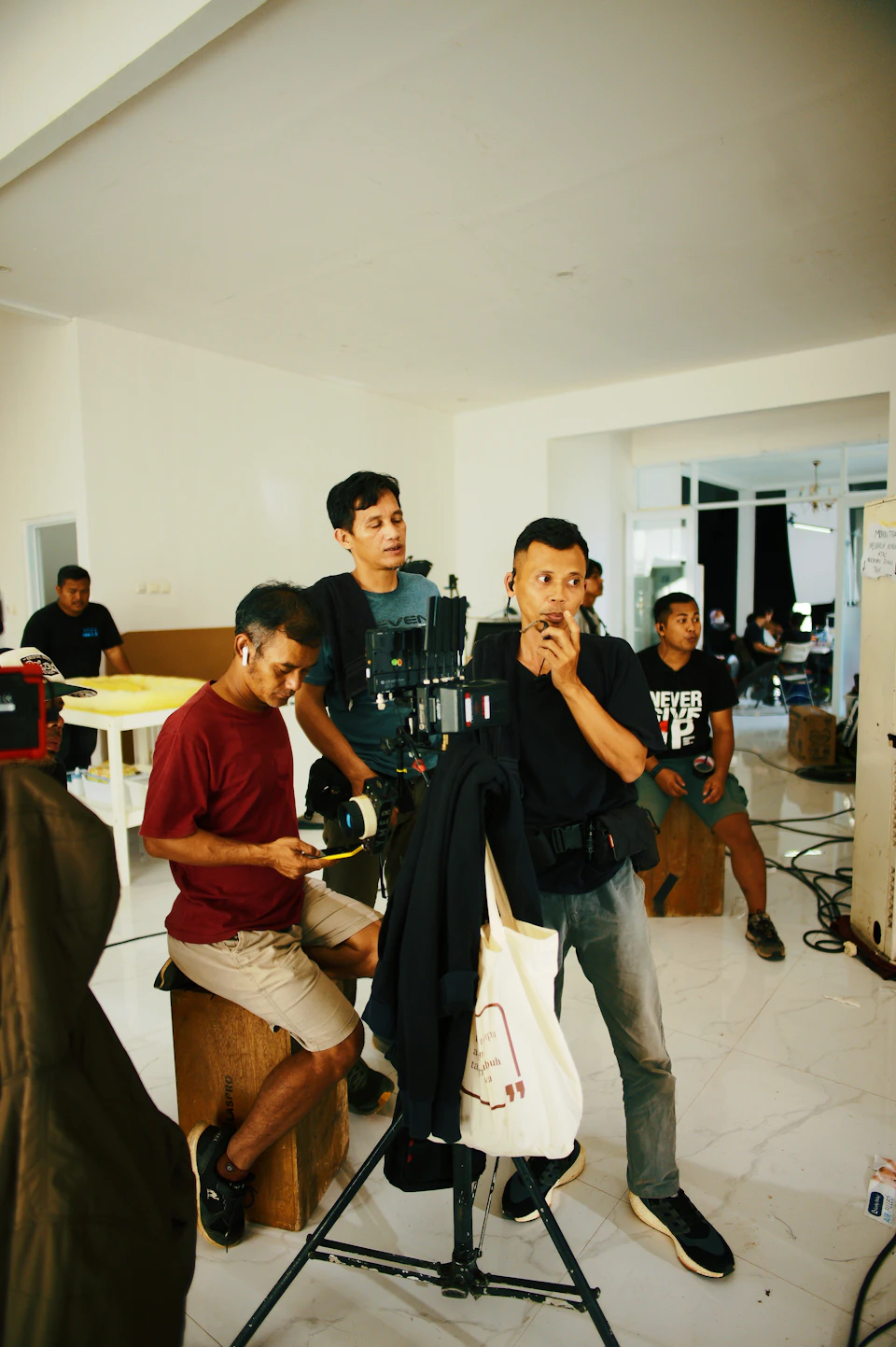 The image depicts a group of people gathered around a camera set up on a tripod, likely for filming purposes. There are four individuals visible in the scene - two men sitting on a bench and two standing behind them. The man seated on the bench is holding a microphone, while another person stands nearby with a camera attached to it. A third individual can be seen standing further back from the group, possibly operating the camera or assisting with the filming process.