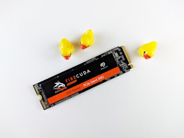 A computer SSD labeled 'FIRECUDA Gaming 520 Series PCIe Gen4 SSD' lies next to three small rubber ducklings with yellow bodies and red beaks, set against a white background.