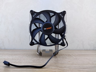 A computer CPU cooler with a large black fan mounted on a reflective heat sink, featuring the brand name in the center. It is situated on a light wooden surface with a power cable extending in front.