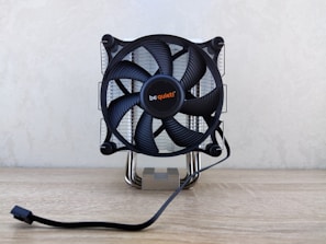 A computer CPU cooler with a large black fan mounted on a reflective heat sink, featuring the brand name in the center. It is situated on a light wooden surface with a power cable extending in front.