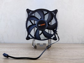 A computer CPU cooler with a large black fan mounted on a reflective heat sink, featuring the brand name in the center. It is situated on a light wooden surface with a power cable extending in front.