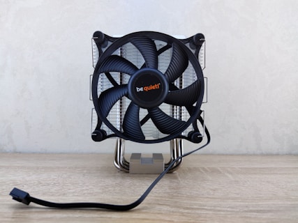 A computer CPU cooler with a large black fan mounted on a reflective heat sink, featuring the brand name in the center. It is situated on a light wooden surface with a power cable extending in front.