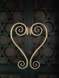 Ornamental metal handles with intricate scroll designs form a symmetrical heart shape against a dark, textured door. The door features geometric patterns creating a decorative lattice look.
