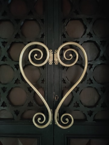 Ornamental metal handles with intricate scroll designs form a symmetrical heart shape against a dark, textured door. The door features geometric patterns creating a decorative lattice look.