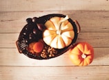 A wicker basket filled with a variety of autumn produce including a white and orange striped pumpkin, dark grapes, dried apricots, and walnuts. An additional small orange pumpkin is placed nearby on a rustic wooden table.