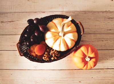 A wicker basket filled with a variety of autumn produce including a white and orange striped pumpkin, dark grapes, dried apricots, and walnuts. An additional small orange pumpkin is placed nearby on a rustic wooden table.