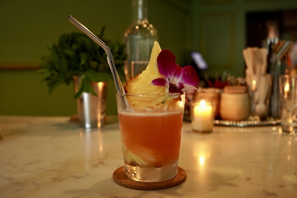 A cocktail glass featuring a vibrant orange beverage, topped with creamy swirl and garnished with a purple orchid flower. A metal straw is inserted into the drink. The background includes a green wall, a small candle, some mint leaves, and an arranged set of bar items such as bottles and glasses.
