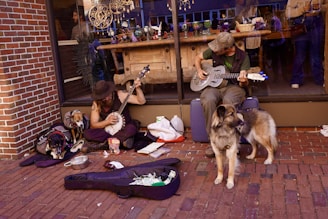 Two musicians are sitting on a sidewalk in front of a brick wall and a large window, playing instruments. One is playing a banjo, and the other is playing a resonator guitar. Both musicians appear relaxed, with a dog sitting nearby and another sleeping under a backpack. Open instrument cases with various items and some change are on the ground in front of them. In the background, a shop window displays various arts and crafts.