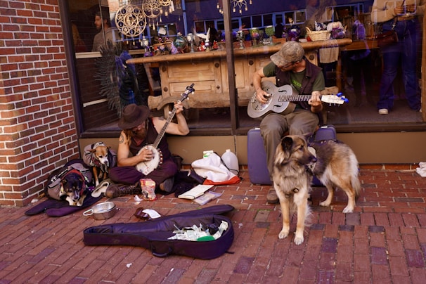 Two musicians are sitting on a sidewalk in front of a brick wall and a large window, playing instruments. One is playing a banjo, and the other is playing a resonator guitar. Both musicians appear relaxed, with a dog sitting nearby and another sleeping under a backpack. Open instrument cases with various items and some change are on the ground in front of them. In the background, a shop window displays various arts and crafts.