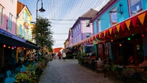 A vibrant street scene featuring colorful buildings on both sides, adorned with festive lights and bunting. The cobblestone path is lined with outdoor seating and plants, creating a lively and welcoming atmosphere. A few people walk down the street, adding a sense of activity and urban energy.