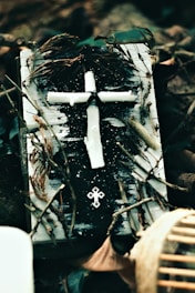 A rustic, handmade cross constructed from what appears to be white candle wax is positioned on a weathered book covered with black and white paint. Dried twigs and leaves encircle the book, contributing to an aged, natural appearance. Another small, metallic cross embellishment rests below the larger cross.