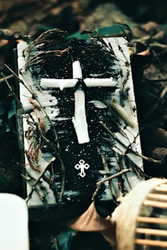 A rustic, handmade cross constructed from what appears to be white candle wax is positioned on a weathered book covered with black and white paint. Dried twigs and leaves encircle the book, contributing to an aged, natural appearance. Another small, metallic cross embellishment rests below the larger cross.