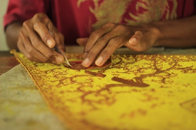 Hands are working on a detailed yellow and brown batik fabric using a small tool. The fabric displays an intricate pattern of branches and leaves, suggesting a traditional craft or textile art activity.