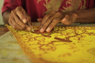Hands are working on a detailed yellow and brown batik fabric using a small tool. The fabric displays an intricate pattern of branches and leaves, suggesting a traditional craft or textile art activity.