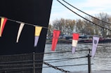 Colorful triangular flags are hanging on a string, with a large dark structure in the background. The setting appears to be near a body of water, with trees and small buildings visible across the water. The flags have various patterns and colors, including yellow, black, white, and red.
